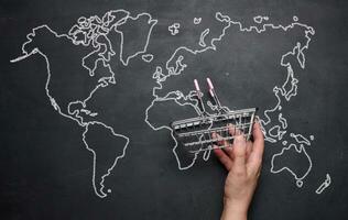 A female hand holding a miniature empty shopping basket against the background of a drawn world map. Concept of online shopping, international shipping photo