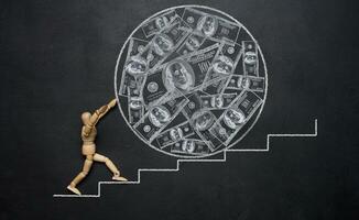 A wooden mannequin rolling a large ball with drawn dollars down the stairs, symbolizing the concept of wealth and hard work towards achieving goals. Inflation and increase in money supply photo