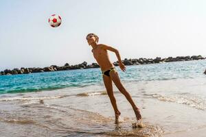 joven chico jugando con un fútbol pelota en el playa por el mar foto