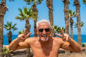 portrait of attractive middle aged man wearing sunglasses making victory sign with canarian palms as background photo