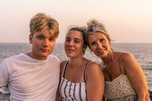 hermosa madre con su dos veinte año antiguo hijos tomar un descanso por el mar a puesta de sol foto