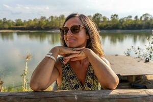 retrato de hermosa medio Envejecido dama vistiendo Gafas de sol en el bancos de el río foto