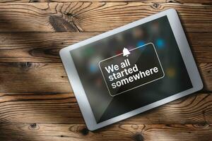 Flat lay photo of digital tablet over wood desk with motivational quote on screen. We all started somewhere quote concept with copy space.