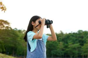 asiático niña usos prismáticos a observar el naturaleza, aves, y animales de el nacional parque mientras cámping con su familia. con el concepto de aprendizaje fuera de el salón de clases naturaleza estudiar, exploración. foto