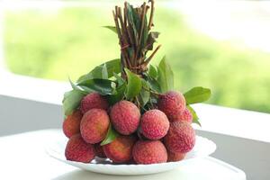 A plate of lychee bunches on a white table, a blurred background of green trees. They are reddish pink or bright red. seed coat Milky white that is full of water. photo