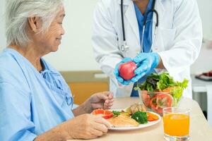 asiático mayor mujer paciente comiendo salmón filete desayuno con vegetal sano comida mientras sentado y hambriento en cama en hospital. foto