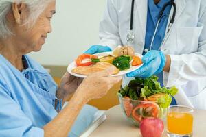 Asian senior woman patient eating Salmon steak breakfast with vegetable healthy food while sitting and hungry on bed in hospital. photo