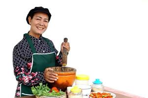 Happy Asian senior woman is cooking, wear chef cap and apron, holds pestle, mortar and plate of chillies, isolated on white background. Concept, Cooking for family. Thai elderly kitchen lifestyle. photo