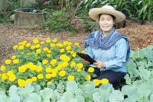 Asian female gardener is at vegetable and flower garden, hold smart tablet, smile. Concept, Using technology wireless to research, control and manage quality of agricultural crop. Smart farmer. photo