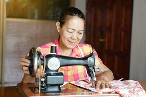 Happy senior Asian woman tailor is sewing cloth by vintage sewing machine at home. Concept, Senior  lifestyle, spend  free time to repair or design clothes. Hobby, not lonely. photo