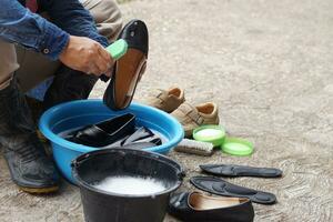 Closeup hands use brush to clean and wash black shoes in black bucket and blue bowl outdoor. Concept, take care, maintenance footwears from dirt and bad smell for using long time. Hygiene and sanitary photo