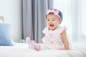 Asian baby toddler in cute pink dress is smiling while sitting on bed with happiness for healthy kid and adorable girl portrait usage photo