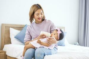 Asian mother is feeding milk from bottle to her cute toddler baby girl in bed for healthy kid nutrition and motherhood by raising child to grow and parenting photo