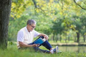 Senior Asian man reading a book while sitting under the tree by the lake at the public park for recreation, leisure and relaxation in nature concept photo