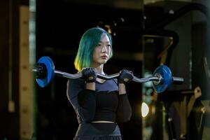 Asian woman is practice weight lifing using easy bar as beginner on barbell for arm and core muscle inside gym with dark background for exercising and workout concept photo