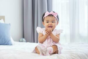 Asian baby toddler in cute pink dress is smiling while sitting on the bed with happiness for healthy kid and adorable girl portrait usage photo