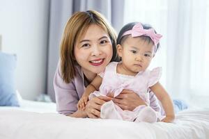 Asian mother is holding her pretty smiling baby daughter while spending quality time in the bed for family happiness and parenting concept photo