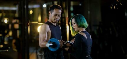 Asian woman is using easy bar as beginner in weight training on barbell for arm and core muscle inside gym with support from trainer to prevent injury dark background for exercising and workout photo