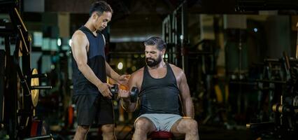 Caucasian man is using dumbbell in weight training for arm muscle inside gym with support from trainer to prevent injury dark background for exercising and workout concept with copy space photo