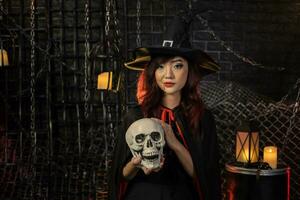 Asian girl in witch costume holding human skull for halloween party trick or treat concept with dark black background photo