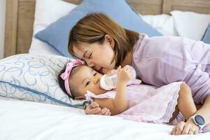 Asian mother is feeding milk from bottle to her cute toddler baby girl in bed for healthy kid nutrition and motherhood by raising child to grow and parenting photo