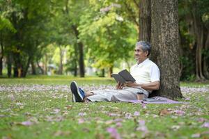 portrait happy senior man sitting against the tree and reading a book in the nature park, concept of senior activities,hobby,relaxing,health benefits in nature photo