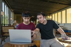 young asian man working remotely on internet online over laptop on vacation while take a break with his friend in cafe on mountain, destination in Thailand.concept of travel freely,digital nomad photo