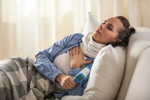 Ill woman experiencing breathing and cough problems while holding forehhead thermometer and lying down on a sofa photo