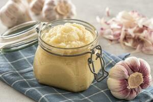 Aromatic garlic paste in a glass jar laid on rustic kitchen cloth with bulbs and peeled cloves photo