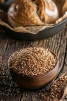 Rural wooden bowl full of wheat grains ready to grind for fresh flour. crunchy yeast bread loaf in the background photo