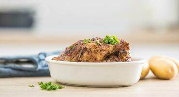 Whole chicken roasted in white dish with raw potatoes on kitched table. Home interior in the background photo