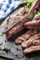 Freshly grilled tomahawk steak on slate plate with salt pepper rosemary and parsley herbs. Chef with knife cuts juicy bovine steak photo