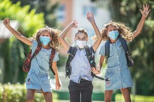 contento Niños de escuela con cara mascaras saltar desde alegría a regreso a colegio durante el covid-19 cuarentena. foto