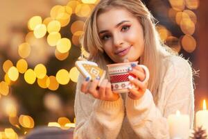 un joven niña en un lana suéter participación un pan de jengibre y un taza con té café o Navidad puñetazo foto