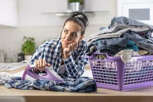 mujer es soñador mientras planchado siguiente a un sobrellenado grande cesta lleno de ropa foto