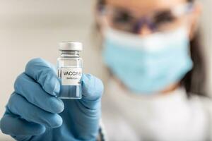 Medical worker wearing protective mask holds a dose of Covid 19 vaccine in her hand towards the camera photo