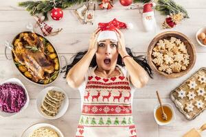 Stressed female cook in a Christmas apron and Santa hat holding her head and lying on the floor surrounded by gingerbread, Linz cakes, on the other side, roast goose or turkey with side dishes photo