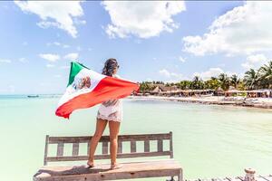 Woman stands on a wooden bench holding Mexican flag looking towards seaside with beach, palms and houses photo