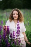 Young woman stands in white shirt in field of purple and pink lupins. Beautiful young woman with curly hair outdoors on a meadow, lupins blossom. Sunset or sunrise, bright evening light photo