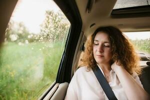 Cheerful positive curly young woman in casual wear sitting in automobile backseat with fastened seatbelt and looking away. photo