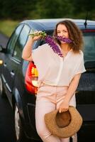 Young woman stands in white shirt near car with purple and pink lupins. Beautiful young woman with curly hair with bouquet of lupins. Sunset or sunrise, bright evening light photo