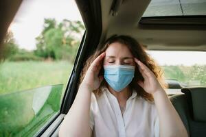 elegante joven carly mujer en médico máscara tener un dolor de cabeza mientras sentado en espalda asiento de coche en natural antecedentes con puesta de sol foto