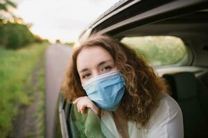elegante joven carly mujer en médico máscara mirando fuera ventana mientras sentado en espalda asiento de coche en borroso antecedentes con puesta de sol foto