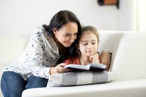 apoyo mujer lee un libro a lo largo con un niña a hogar en un sofá foto