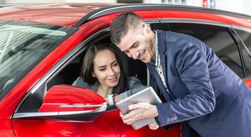 Car dealership employee with tablet shows woman intending to buy a red car she sits in technical features photo