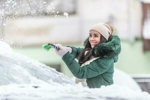 Young female in winter clothes cleans car from snow outside the apartment block in the winter photo