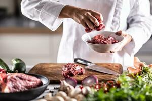 Detail of red meat chunks put into a white bowl on a meal preparation table full of ingedients photo