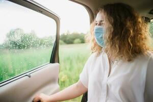 elegante joven carly mujer en médico máscara mirando fuera ventana mientras sentado en espalda asiento de coche en borroso antecedentes con puesta de sol foto