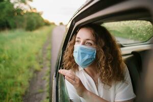 elegante joven carly mujer en médico máscara mirando fuera ventana mientras sentado en espalda asiento de coche en borroso antecedentes con puesta de sol foto