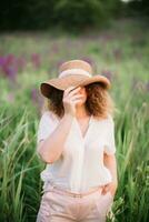 Young woman stands in white shirt in field of purple and pink lupins. Beautiful young woman with curly hair and hat outdoors on a meadow, lupins blossom. Sunset or sunrise, bright evening light photo